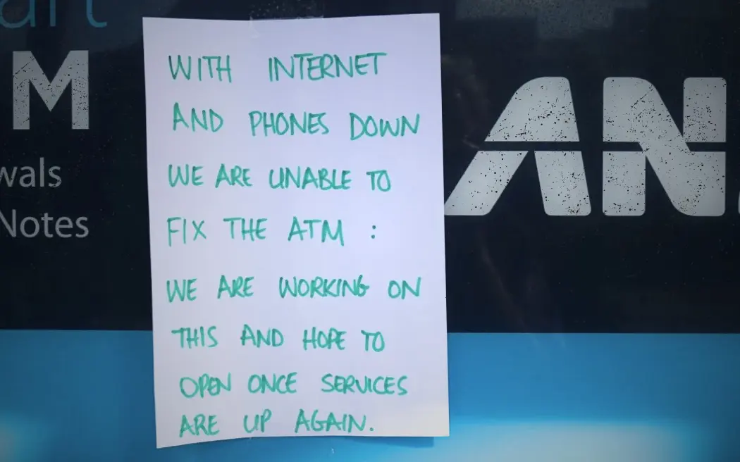 Handwritten sign taped to an ANZ ATM reading: "With internet and phones down we are unable to fix the ATM. We are working on this and hope to open once services are up again." The ANZ logo and partial ATM screen are visible behind the sign.