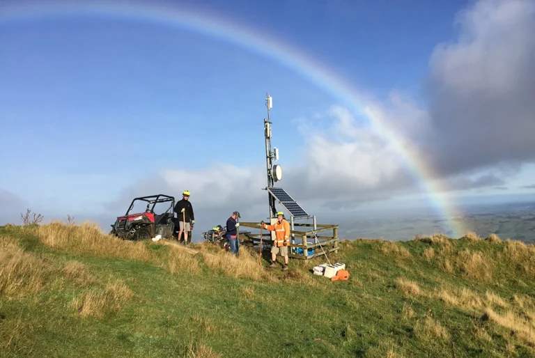 Three technicians working on a solar-powered rural wireless tower on a hilltop in the Ruahine ranges, New Zealand, with a full rainbow arching overhead and farmland visible in the valley below